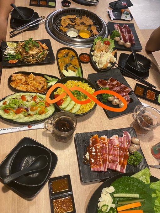 A Korean BBQ meal spread on a wooden table features grilled meat on a tabletop grill, raw marinated meats, rice, salad, dipping sauces, and a red infinity-like decoration.