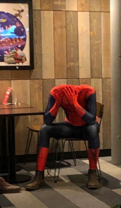 A person in a Spider-Man costume sitting on a chair at a table with their head in their hands, appearing dejected, with a framed painting visible on the wooden wall behind them.