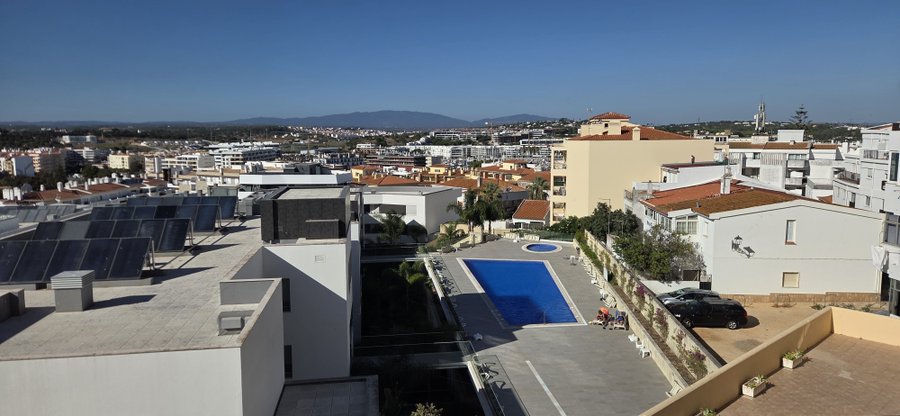 A sunny rooftop view overlooks a coastal town's residential area with white buildings, terracotta roofs, a swimming pool, and distant hills or mountains under a clear blue sky.
