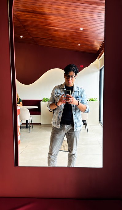A man with glasses takes a mirror selfie while wearing a denim jacket in a room with a curved ceiling.