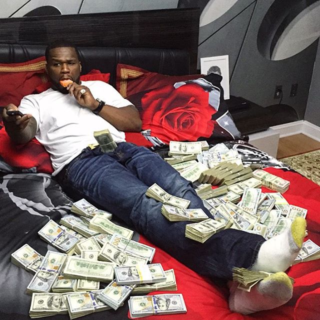 A man lies on a rose-patterned bed casually surrounded by scattered and stacked bundles of U.S. $100 bills, looking at his phone. The "Crypto holders in a few months" caption satirizes investor optimism by juxtaposing the lavish wealth depicted with the often unrealistic expectation that crypto profits will make users this rich.