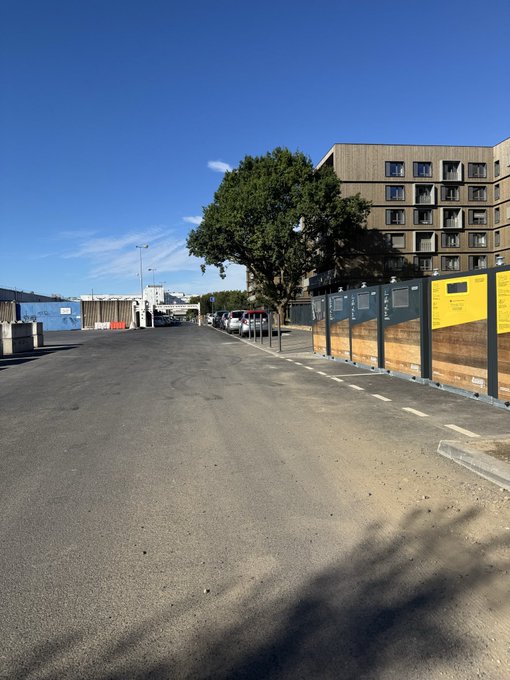 An outdoor scene showing an empty asphalt road or parking area bordered by a wooden fence with yellow signs, with a multi-story beige apartment building and trees under a clear blue sky.