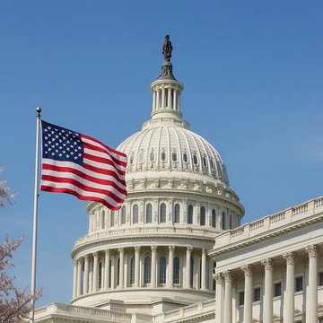 The U.S. Capitol Building with its white dome and the Statue of Freedom on the roof, with an American flag waving in the foreground against a clear blue sky.
