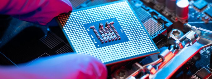 A gloved hand installs a processor onto a motherboard, with visible pins and electronic components, visually reinforcing the post's theme of AI chip demand outstripping supply and the need for better orchestration of hardware resources like those provided by io.net.