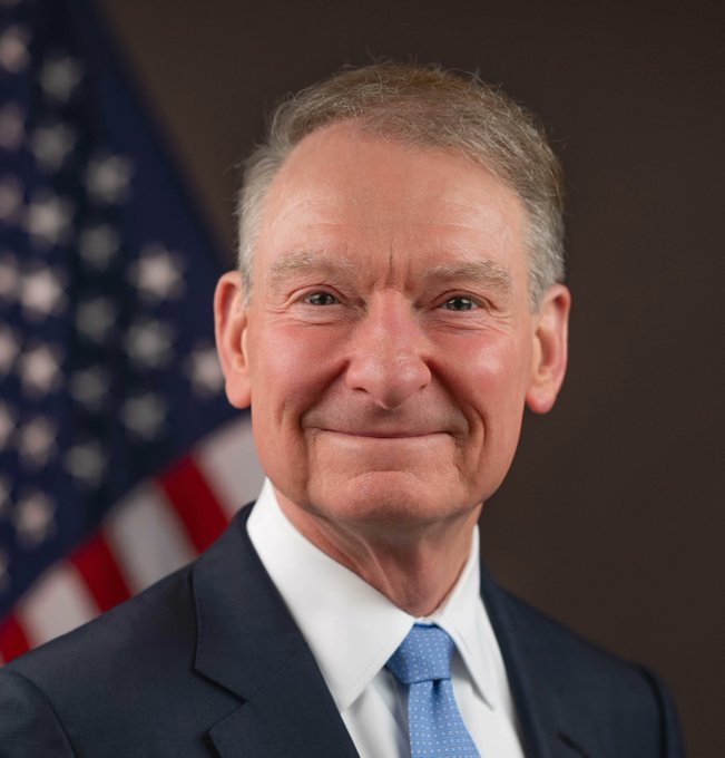 A formal portrait shows an older man with short gray hair, wearing a dark suit, white shirt, and light blue tie, smiling in front of an American flag. The image relates to the post by depicting a figure in official political/governmental capacity, suggesting connection to SEC crypto regulation developments.