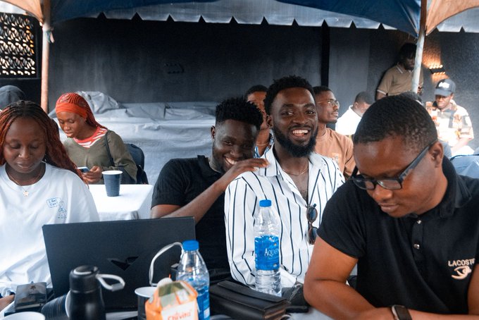 A photograph of a group of people, with three men in the foreground smiling, seated at a table with water bottles and a laptop under a tent.