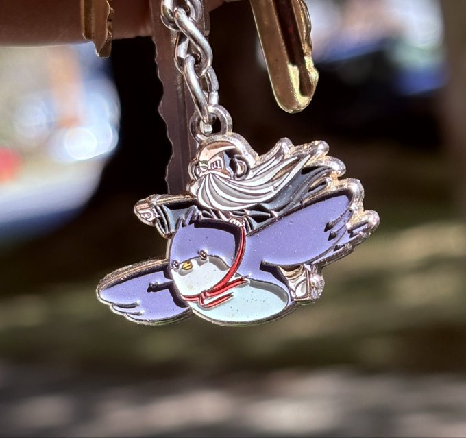 A silver keychain charm features an enameled design of a cloaked wizard riding a shark-like creature against a blurred outdoor background.