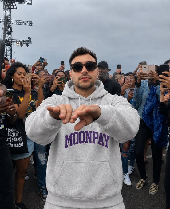 A man wearing sunglasses and a gray "MOONPAY" hoodie gestures toward the camera while surrounded by a crowd of fans holding up their phones at an outdoor event.