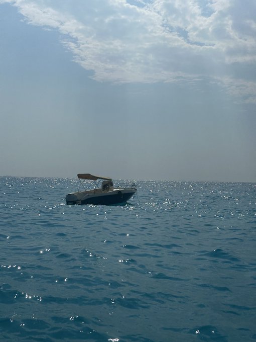 A small motorboat floats alone on a sparkling blue sea under a hazy sky.