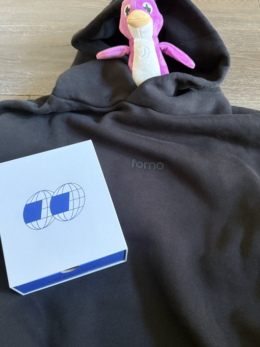 A black hoodie with the word 'fomo' embroidered on the chest is laid out with a purple and white penguin plush peeking out from the hood. A white box featuring a blue logo of two interlocking globes sits on top of the garment.