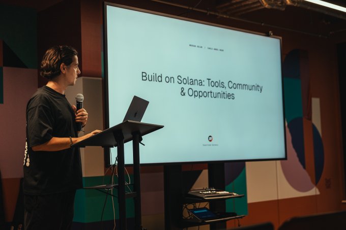 A person stands at a podium with a microphone in front of a screen displaying the title slide 'Build on Solana: Tools, Community & Opportunities' along with 'WORKSHOP | COINGECKO' at the top and the CoinGecko logo.