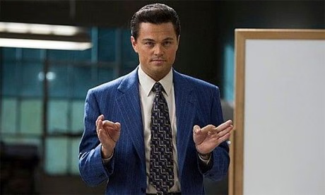 Leonardo DiCaprio as Jordan Belfort in The Wolf of Wall Street, wearing a blue pinstripe suit and patterned tie, making a 'money' gesture with his hands.