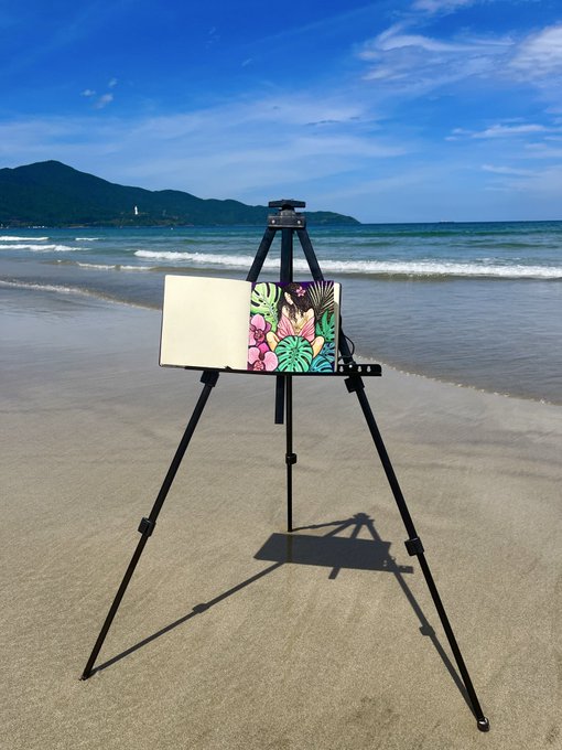 A black tripod easel stands on a sandy beach holding a sketchbook and a small tropical-themed painting, with ocean waves and distant mountains visible under a blue sky.
