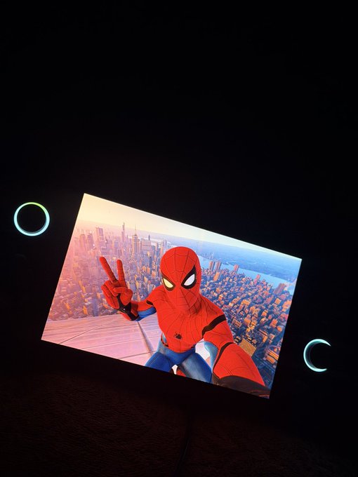 A photo of a glowing rectangular display or light panel showing a scene of the superhero standing on a rooftop overlooking a city, with two illuminated circular buttons or speakers visible on the sides against a dark background.