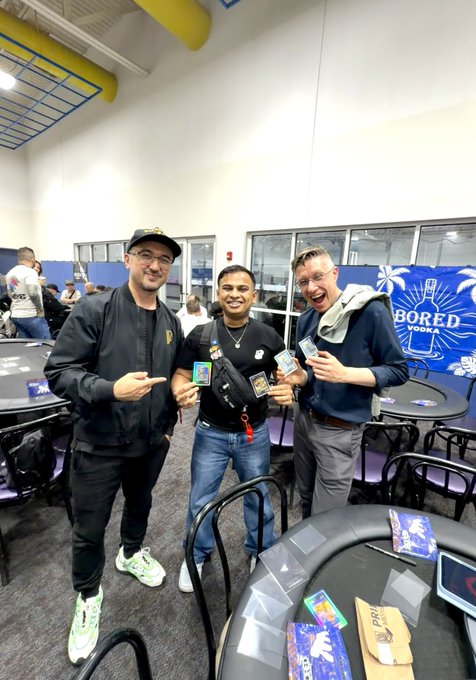Three men posing indoors at what appears to be a 'BORED VODKA' event, holding branded merchandise and smiling, with a blue branded backdrop visible behind them.