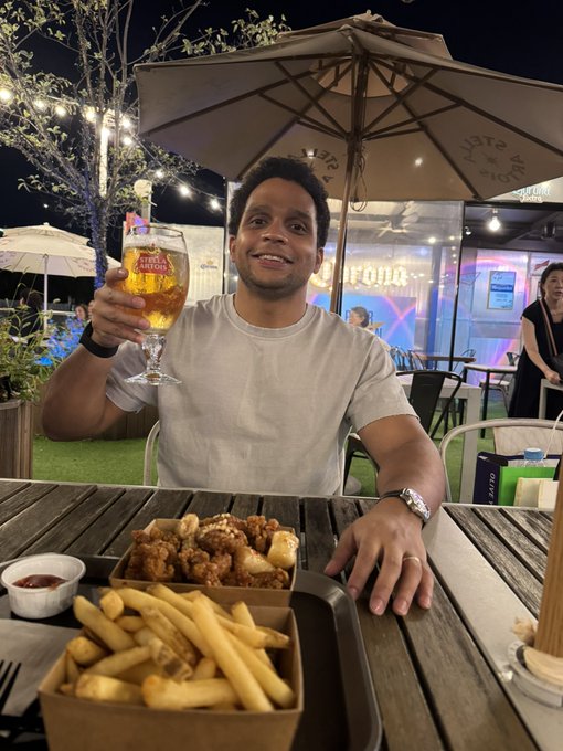 A man wearing a beige t-shirt sits at an outdoor wooden table smiling and holding up a glass of beer, with a basket of fries and fried chicken in front of him and string lights and umbrellas in the background.