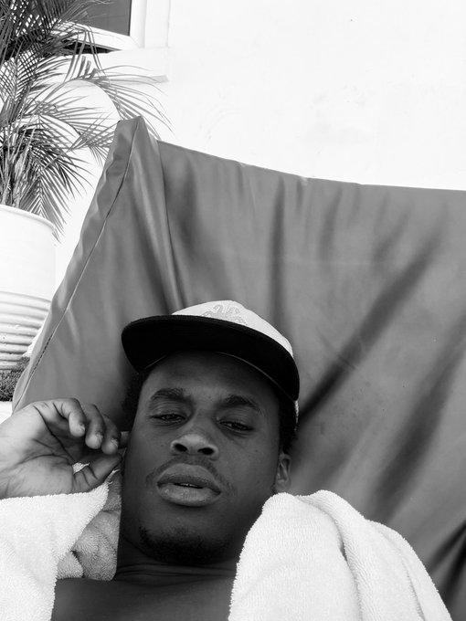A relaxed man wearing a cap with visible "DADA" text lounges on a sunbed with a towel draped over his shoulders, embodying the laid-back vibe of a Nigerian web3 founder enjoying downtime. The casual selfie reflects his self-described role as the brain behind @l0f0_00, reaching out to connect with the onchain community.