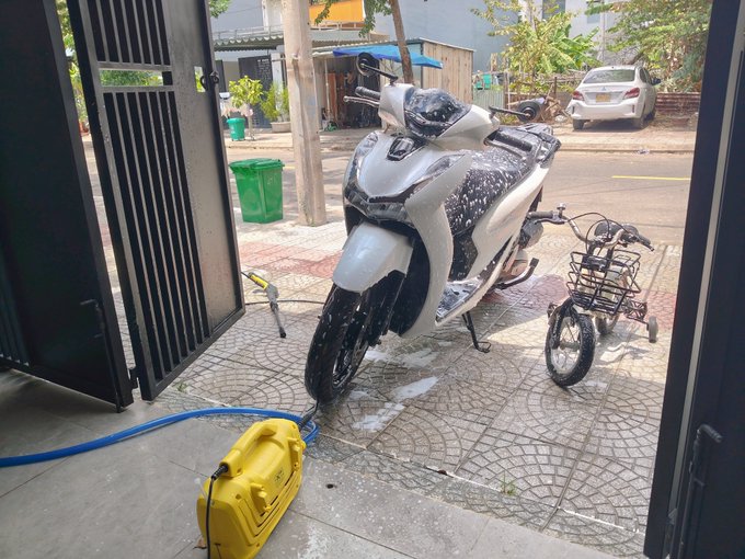 A white motor scooter covered in soap suds parked on a tiled sidewalk next to a yellow pressure washer unit, with a small bicycle nearby.
