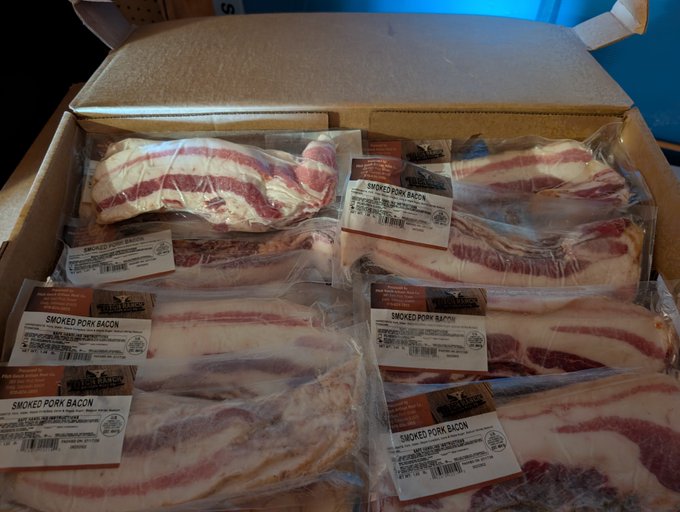 A cardboard box filled with vacuum-sealed packages of smoked pork bacon, each labeled with a nutritional information sticker.