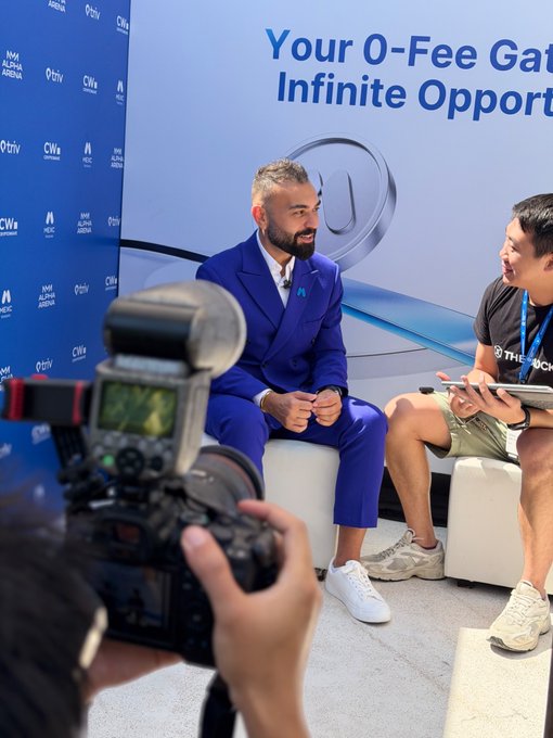 A bearded man in a blue suit is being interviewed at Coinfest Asia, with the "Your 0-Fee Gate to Infinite Opport..." banner behind him, while a photographer captures the moment—embodying the event's mix of professional networking and energetic atmosphere highlighted in the post.