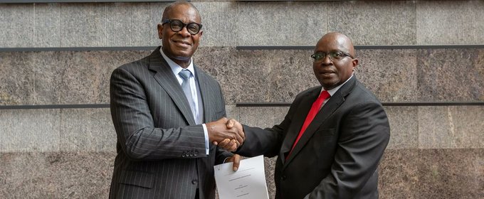 Two men in business suits shaking hands and exchanging a document, one wearing a red tie, posing in front of a stone wall.