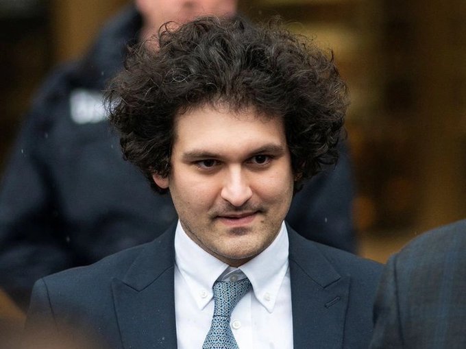 The image shows a man with curly hair wearing a suit and tie standing next to another person who appears to be a police officer. The man has his head turned towards the camera while looking at it. The background of the photo features a building, suggesting that they might be in an urban setting or court room.