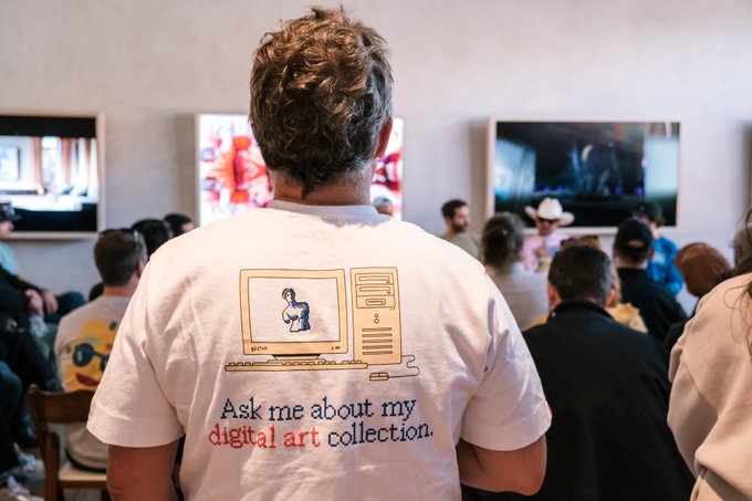 A person viewed from behind attends an art event, wearing a white t-shirt featuring a graphic of a computer and the text 'Ask me about my digital art collection.'