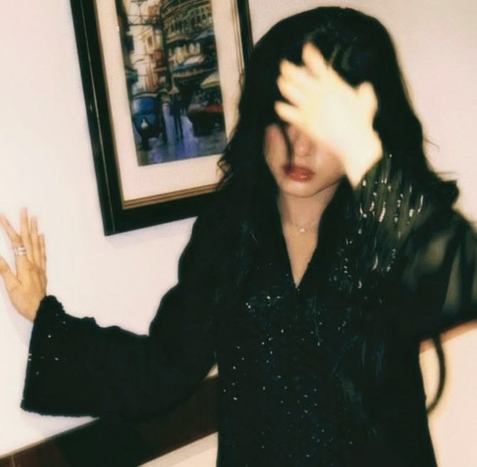 A vintage-style photograph of a woman with long dark hair wearing a black sequined top, covering her face with her hand as she stands beside a framed picture.