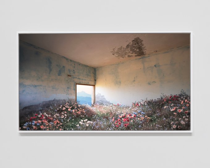 A framed photograph depicting an abandoned room with peeling walls where wildflowers have overgrown the floor, with a window showing a mountain view.