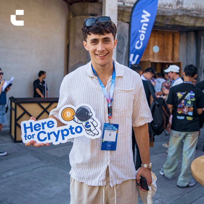 A smiling young man at an outdoor event holding a sign that reads 'Here for Crypto' with a Bitcoin astronaut graphic, wearing a lanyard with a festival pass and sunglasses on his head, with a 'CoinW' banner visible in the background.