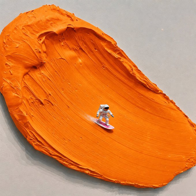 An artistic composition featuring a tiny astronaut surfing on a surfboard through a large smear of thick orange paint on a gray surface.
