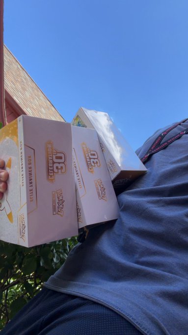 Two 'Celebration Box' packages containing '30 Hits' and '50 Hits' cards are held outdoors against a gray shirt and blue sky.