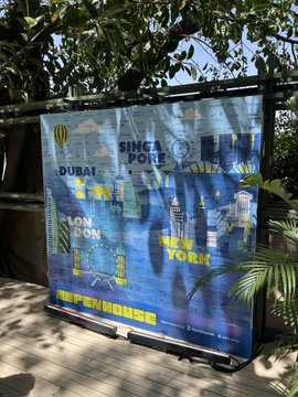 An outdoor banner displaying world city names (Dubai, Singapore, Hong Kong, New York, London, Sydney) over a blue background, partially shaded by tree leaves.