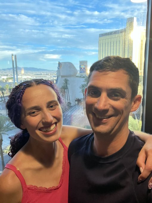 A smiling couple takes a selfie together, with the Mandalay Bay hotel and other Las Vegas Strip buildings visible through the window behind them.