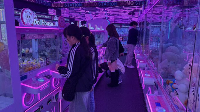 An arcade scene with people playing games at claw machines in a neon-lit environment, featuring a 'Doll House' sign in pink lighting.