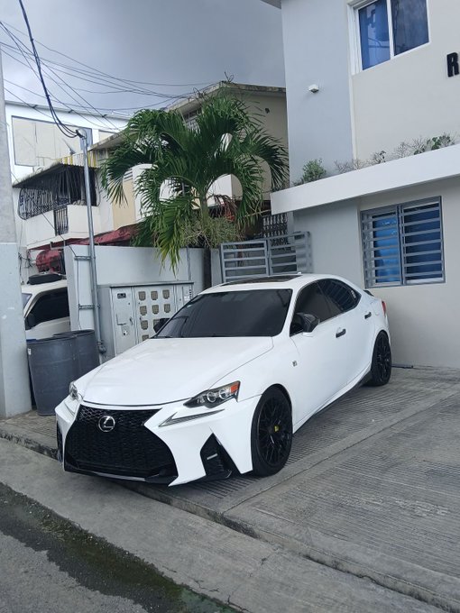 A white Lexus coupe is parked on a street in a residential area, with another similar white car visible behind a fence.