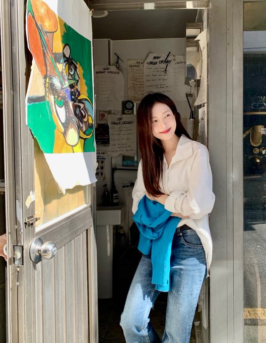 A young woman with long dark hair stands in a doorway holding a blue garment, wearing a white shirt and blue jeans, with colorful artwork visible on the door beside her.