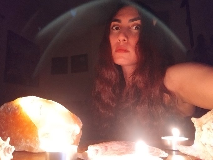 A woman with long red hair sits in a dimly lit room surrounded by glowing crystals, candles, and seashells in what appears to be a mystical or ritualistic setting.