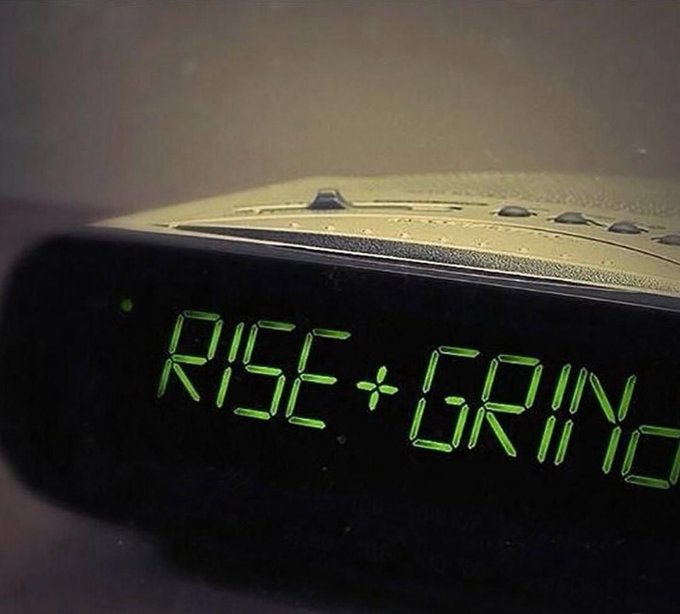 A digital alarm clock displays the words "RISE + GRIND" in glowing green letters, paired with the post's motivational message encouraging followers to have a productive day.