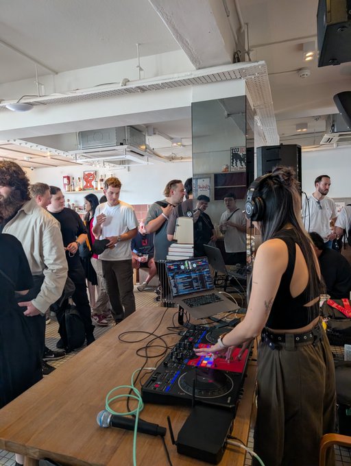 A DJ wearing headphones performs on Pioneer decks and a MacBook setup at a long table, while a crowd gathers to watch and dance in a spacious industrial-style venue, capturing the energetic atmosphere of the Starknet Coffee Rave.