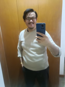 A man with glasses smiles as he takes a mirror selfie with a smartphone, wearing a cream-colored sweater and dark pants.