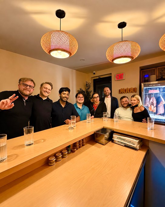 A group of seven people pose behind a wooden bar counter with pendant lights hanging above and a refrigerated meat case visible in the background.