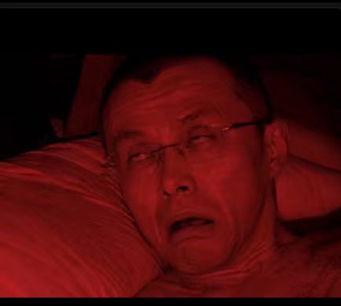 An older man wearing glasses lies in bed, his face illuminated by a red light, appearing distressed or crying.