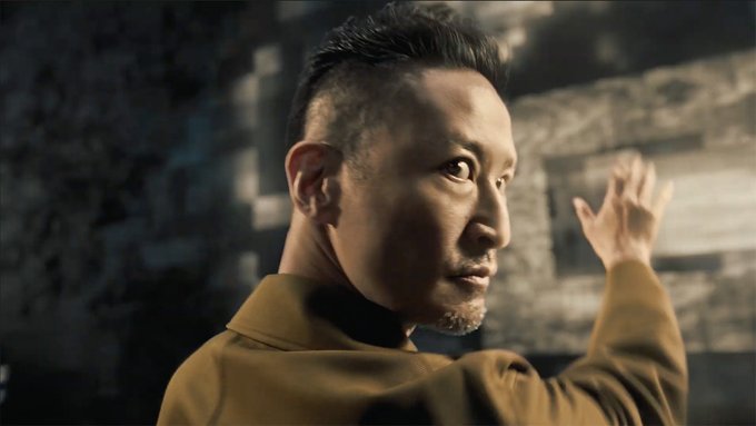 A still shows a man with short dark hair and a goatee, wearing a mustard-yellow jacket, looking back over his shoulder with a serious expression against a blurred stone background.

The image relates to the post by featuring 松岡昌宏 in what appears to be a promotional scene for his Binance Japan commercial.