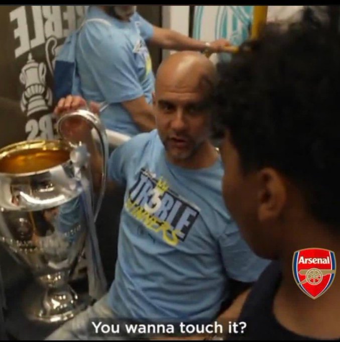 The image features a man wearing a blue shirt holding a trophy with a caption that reads "You wanna touch it?". The trophy appears to be made of metal, possibly silver or gold, and has an inscription on it. The background shows two other people standing in front of a wall adorned with various logos.
