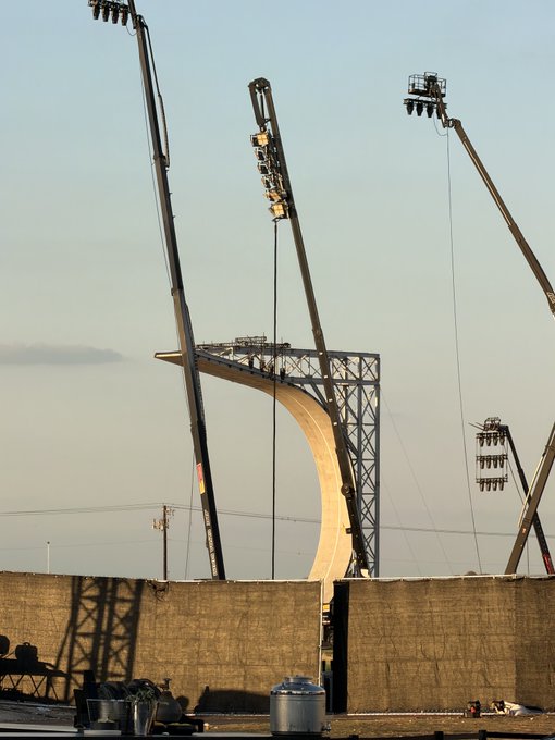 A construction scene at sunset or sunrise showing cranes working on a large concrete structure, likely a stadium or arch.