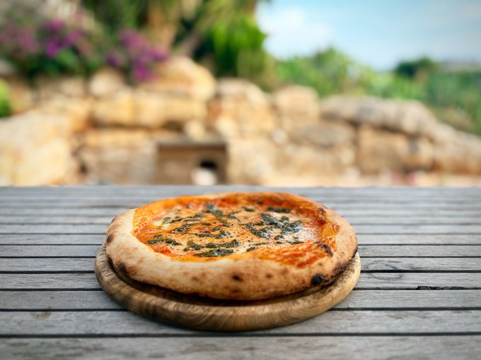 A pizza topped with vibrant green pistachio pesto sits on a wooden board, its colorful, splattered appearance mirroring a Jackson Pollock painting. The artisanal pie rests on a rustic wooden table with a blurred natural backdrop.