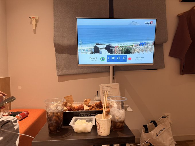 A photo of a living room scene where a TV broadcasts a Brazil vs Japan soccer match, with takeout food, drinks, and snacks spread across a coffee table in the foreground.