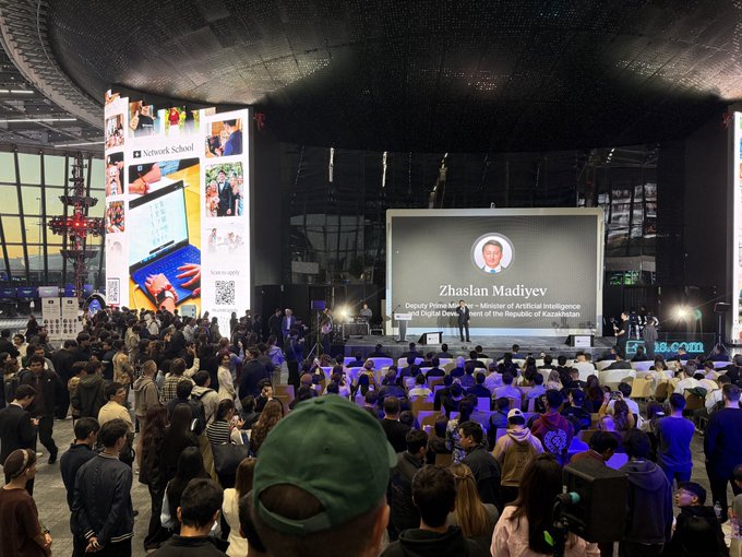 A crowded conference hall with attendees seated facing a large screen displaying 'Zhaslan Madiyev, Deputy Prime Minister - Minister of Artificial Intelligence and Digital Development of the Republic of Kazakhstan', with a Network School booth visible to the left.