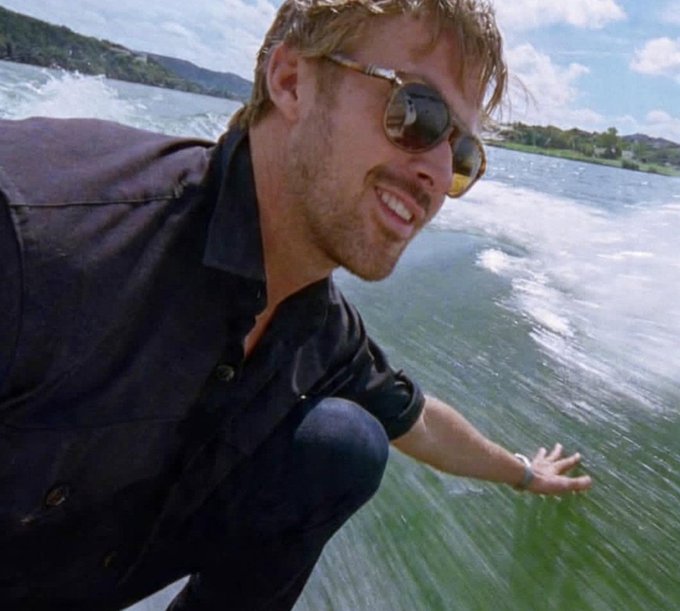 A selfie of a smiling man with blonde hair and sunglasses, wearing a dark shirt, riding a boat across a green lake with a wake trailing behind.